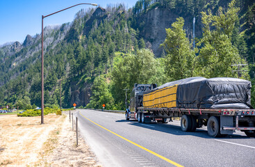 Powerful dark big rig semi truck tractor transporting covered and fastened cargo on flat bed semi...