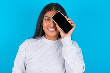 Young hispanic woman wearing grey sweatshirt over blue background holding modern smartphone covering one eye while smiling
