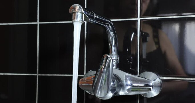 Mature Woman Turning Kitchen Faucet On And Off, Fresh Water Coming Out Of Faucet, Reflected On Black Tile Wall And Faucet Metal, Drinking Water, Female Hands