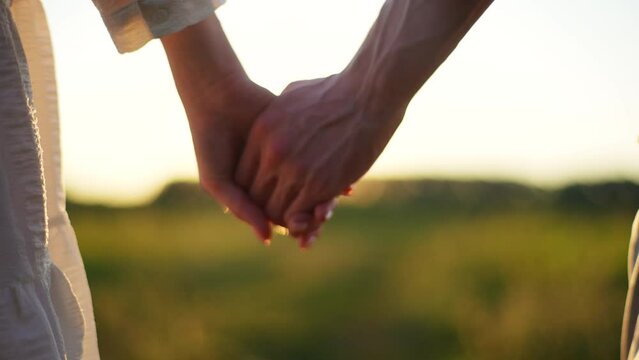 Close-up of unrecognizable young couple in love taking each other hand and walking across green field towards sunset on beautiful summer evening. Wonderful romantic moment between happy couple.