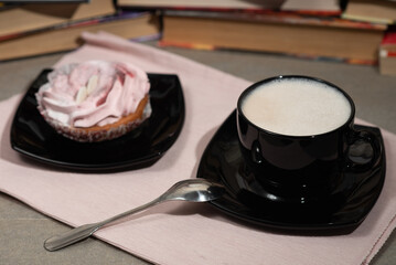 soft juicy biscuit cake of pink color with white cream and a cup with coffee and a pattern of cinnamon on milk foam in a black bowl close-up on a light background