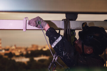 Industrial mountaineering worker in uniform hangs over building and paint roller construction at dusk urban background, painting facade. Rope access laborer on house. Industry night city work concept