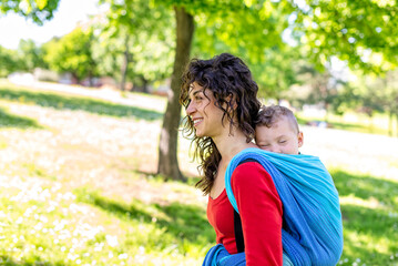 Fototapeta premium close up portrait of a happy young mom carries a child on her back tied up with a colorful traditional piece of fabric in a public park garden. mother and son in a baby wrap sling chilling outdoor.