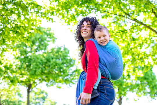 Happy Young Mom Carries A Child On Her Back, Tied Up With A Colorful Traditional Piece Of Fabric In A Public Park Garden. Mother And Son In A Baby Wrap Sling Chilling Outdoor. Family And Togetherness