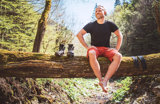 Smiling middle-aged man sitting on the fallen tree log over the mountain forest stream with his beagle dog while he waiting for laundry drying and trekking boots. Traveling with pets concept image.