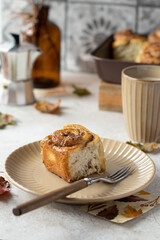 Cinnamon bun on a beige ceramic plate nearly fork with wooden handle.  Tiles on background. Baking dish, mug, vase with dry pine branch, geyser coffee maker and maple leaves on a table. Autumn mood