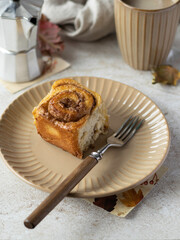 Cinnamon bun on a beige ceramic plate nearly fork with wooden handle.  Tiles on background. Baking dish, mug, vase with dry pine branch, geyser coffee maker and maple leaves on a table. Autumn mood