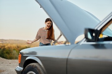 A sad woman has opened the hood of a broken down car and is looking for the cause of the breakdown on a road trip alone with her hands spread apart and shouting in anger