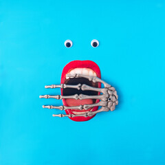 A wide open mouth with red lipstick, white plastic eyes and a skeleton hand over the mouth. Blue background. Surreal minimal horror concept for Halloween celebration banner or card