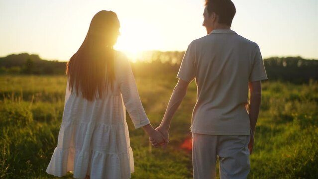 Rear view of happy young couple in love taking each other hand and walking across field towards sunset on beautiful summer evening. Back view of loving man and woman hugging on background of sunlight.
