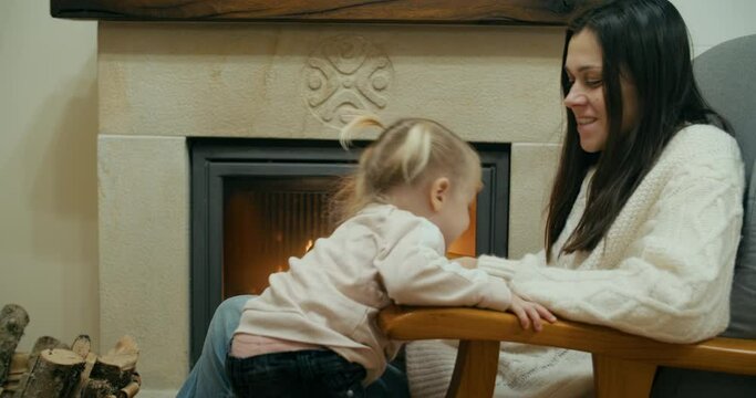 Woman enjoy watching burning fireplace on winter vacation at apartment her cheerful daughter comes to hug mother. Cute children kiss mom by the chimney. Burning fire in indoor fireplace. 