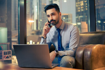 Handsome Indian man talking on mobile phone and using laptop computer at office.