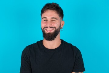 Fototapeta premium Coquettish young hispanic bearded man wearing black T-shirt standing against blue background smiling happily, blinking at camera in a playful manner, flirting with you.