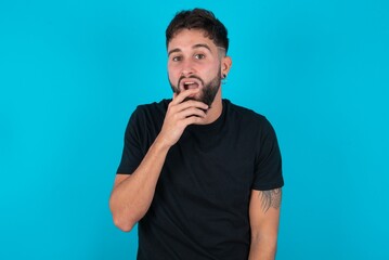 Nervous puzzled young hispanic bearded man wearing black T-shirt standing against blue background opens mouth from surprise, reacts on sudden news.