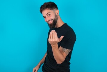 young hispanic bearded man wearing black T-shirt standing against blue background inviting to come with hand. Happy that you came - obrazy, fototapety, plakaty