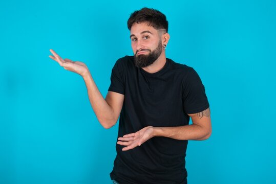 young hispanic bearded man wearing black T-shirt standing against blue background pointing aside with both hands showing something strange and saying: I don't know what is this. Advertisement concept.