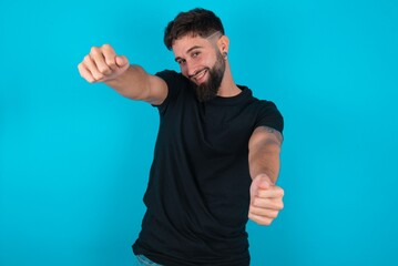 young hispanic bearded man wearing black T-shirt standing against blue background imagine steering wheel helm rudder passing driving exam good mood fast speed