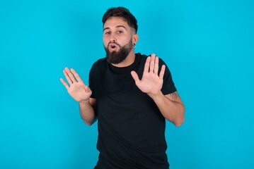 young hispanic bearded man wearing black T-shirt standing against blue background Moving away hands palms showing refusal and denial with afraid and disgusting expression. Stop and forbidden.