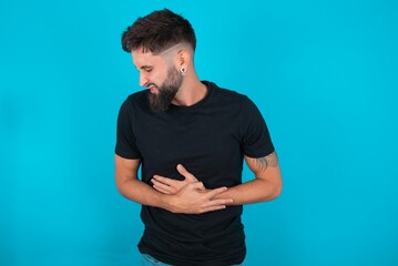 young bearded hispanic man wearing black T-shirt over blue background with hand on stomach because...