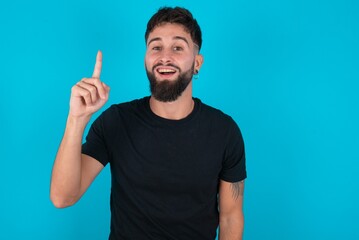 young bearded hispanic man wearing black T-shirt over blue background pointing finger up and...