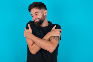 young bearded hispanic man wearing black T-shirt over blue background Hugging oneself happy and positive, smiling confident. Self love and self care