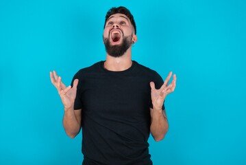 Fototapeta premium young bearded hispanic man wearing black T-shirt over blue background crazy and mad shouting and yelling with aggressive expression and arms raised. Frustration concept.