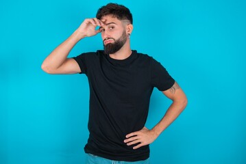 young bearded hispanic man wearing black T-shirt over blue background having problems, worried and stressed holds hand on forehead. - obrazy, fototapety, plakaty