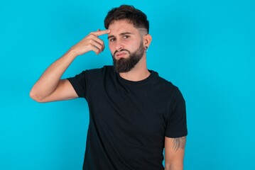 young bearded hispanic man wearing black T-shirt over blue background pointing unhappy at pimple on forehead, blackhead  infection. Skincare concept.