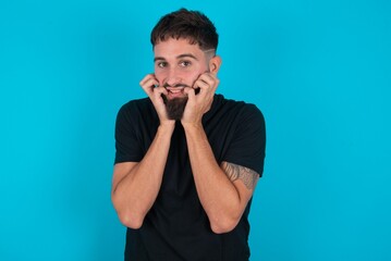 Fearful young bearded hispanic man wearing black T-shirt over blue background keeps hands near mouth, feels frightened and scared,  has a phobia,  Shock and frighted concept.