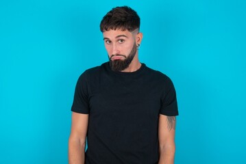 Displeased upset young bearded hispanic man wearing black T-shirt over blue wall frowns face as...
