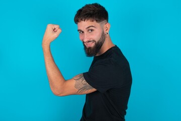 Portrait of powerful cheerful young bearded hispanic man wearing black T-shirt over blue background...