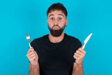 hungry young bearded hispanic man wearing black T-shirt over blue background holding in hand fork...