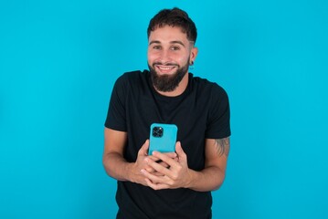 Excited young bearded hispanic man wearing black T-shirt over blue background holding smartphone and looking amazed to the camera after receiving good news.