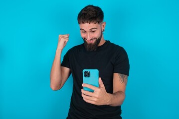 young bearded hispanic man wearing black T-shirt over blue background holding in hands cell and rising his fist up being excited after reading good news.