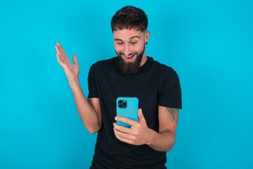 young bearded hispanic man wearing black T-shirt over blue background holding in hands cell reading browsing news