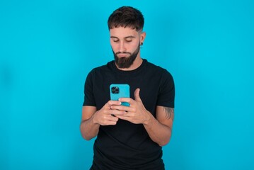 Excited young bearded hispanic man wearing black T-shirt over blue background  winking and eye hold smart phone use read social network news