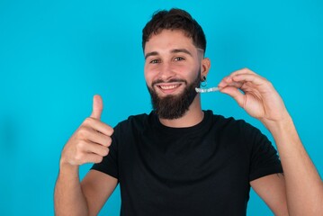 young bearded hispanic man wearing black T-shirt over blue background holding an invisible braces...