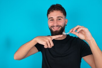 young bearded hispanic man wearing black T-shirt over blue background holding an invisible aligner...