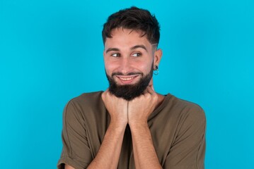young bearded hispanic man wearing casual clothes over blue background holds hands under chin, glad to hear heartwarming words from stranger