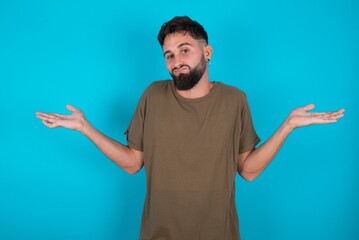 Careless attractive young bearded hispanic man wearing casual clothes over blue background shrugging shoulders, oops.