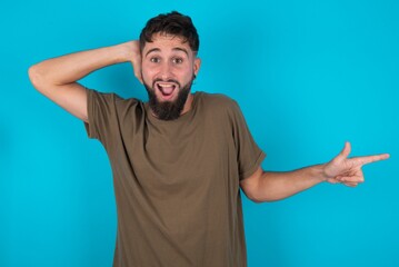 Surprised young bearded hispanic man wearing casual clothes over blue background pointing at empty space holding hand on head