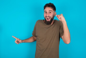 young bearded hispanic man wearing casual clothes over blue background points at empty space holding fist up, winner gesture.