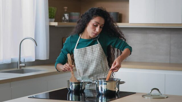 Busy arabian mom attractive curly housewife chef wears apron cooking breakfast at home kitchen stirring meal in saucepans with spoon preparing food for family talking mobile phone cell communication