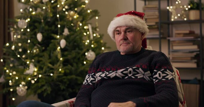 Retired man with gray hair sitting on chair near the Christmas tree. He is wearing warm holiday sweater and Santa hat. The man nervously took off hat, because he is not in the mood to celebrate.