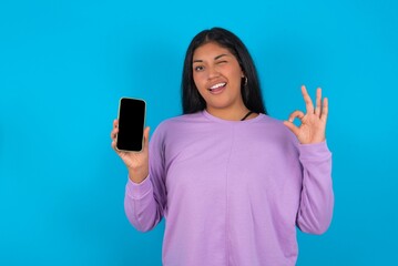 Excited young beautiful latin woman wearing casual clothes standing against blue background showing smartphone blank screen, blinking eye and doing ok sign with hand.  Advertisement concept.