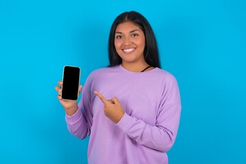 Naklejka premium Smiling young beautiful latin woman wearing casual clothes standing against blue background showing and pointing at empty phone screen. Advertisement and communication concept.