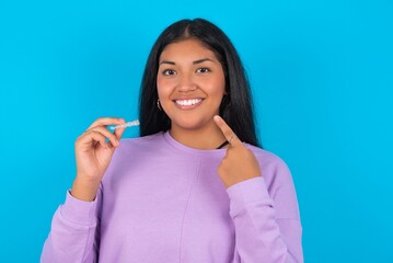 young beautiful latin woman wearing casual clothes standing against blue background holding an invisible aligner and pointing to her perfect straight teeth. Dental healthcare and confidence concept.