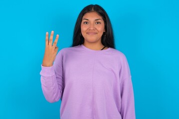 Young beautiful hispanic woman wearing casual clothes over blue background smiling and looking friendly, showing number three or third with hand forward, counting down