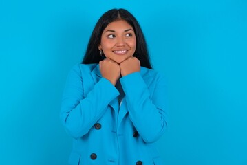 Young latin businesswoman wearing blue blazer over blue background holds hands under chin, glad to hear heartwarming words from stranger