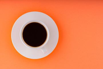 Cup of coffee on orange background. White cup of black coffee on a white saucer. Copy space. Top view